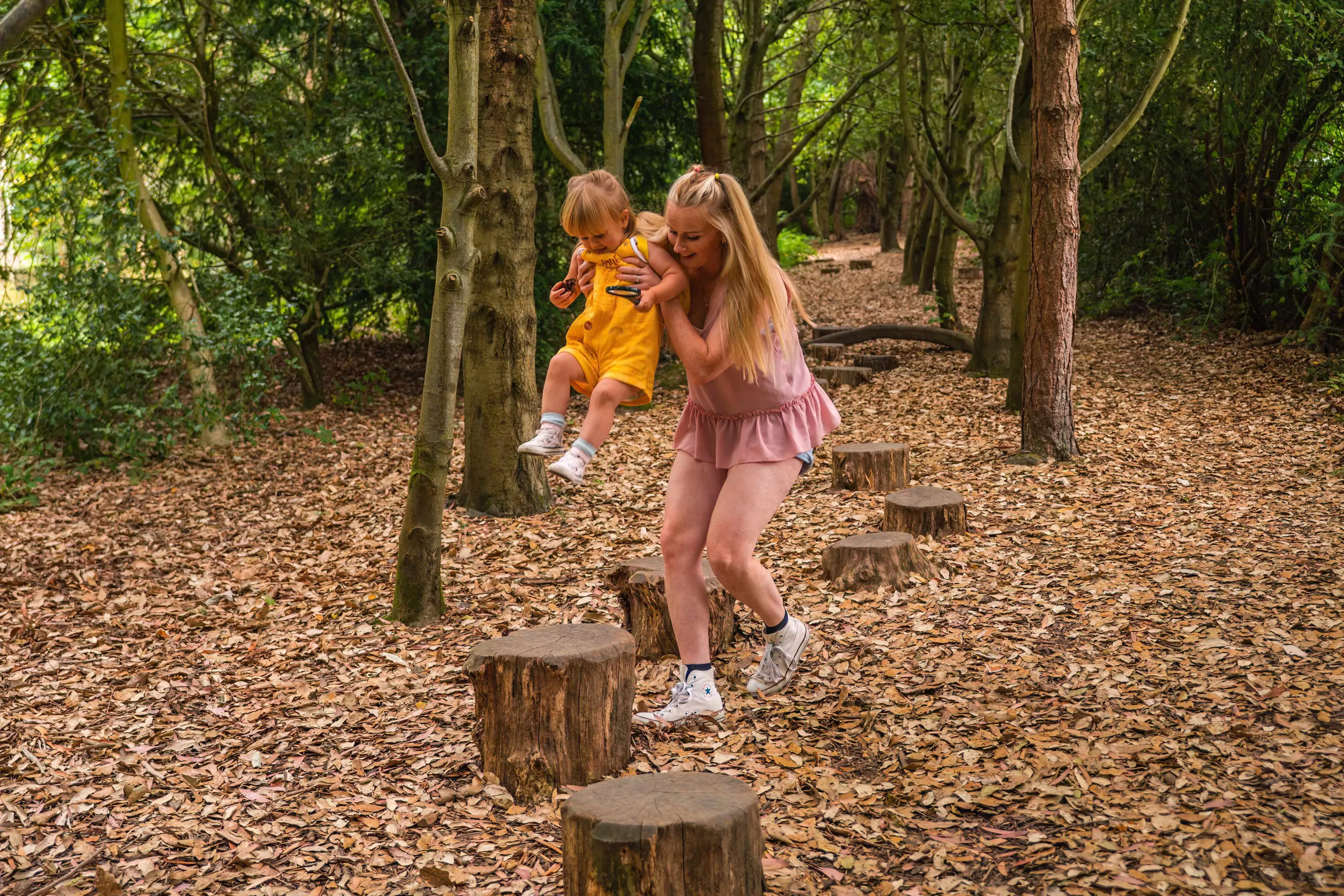 Tree Trunk Trek at Wakehurst, Visual Air © RBG Kew A blonde mother swings a toddler child along a line of tree stumps small enough to use as stepping stones