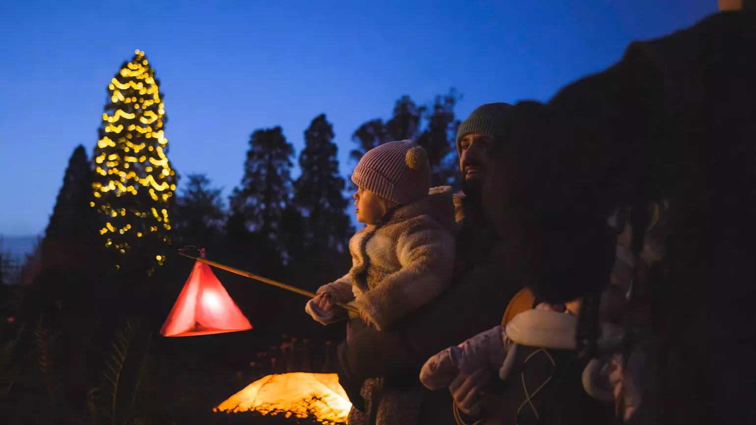 A young family enjoys exploring Glow Wild with a lantern A woman holds a baby, who in turn is holding a lantern and looking towards Christmas lights, which are out of shot