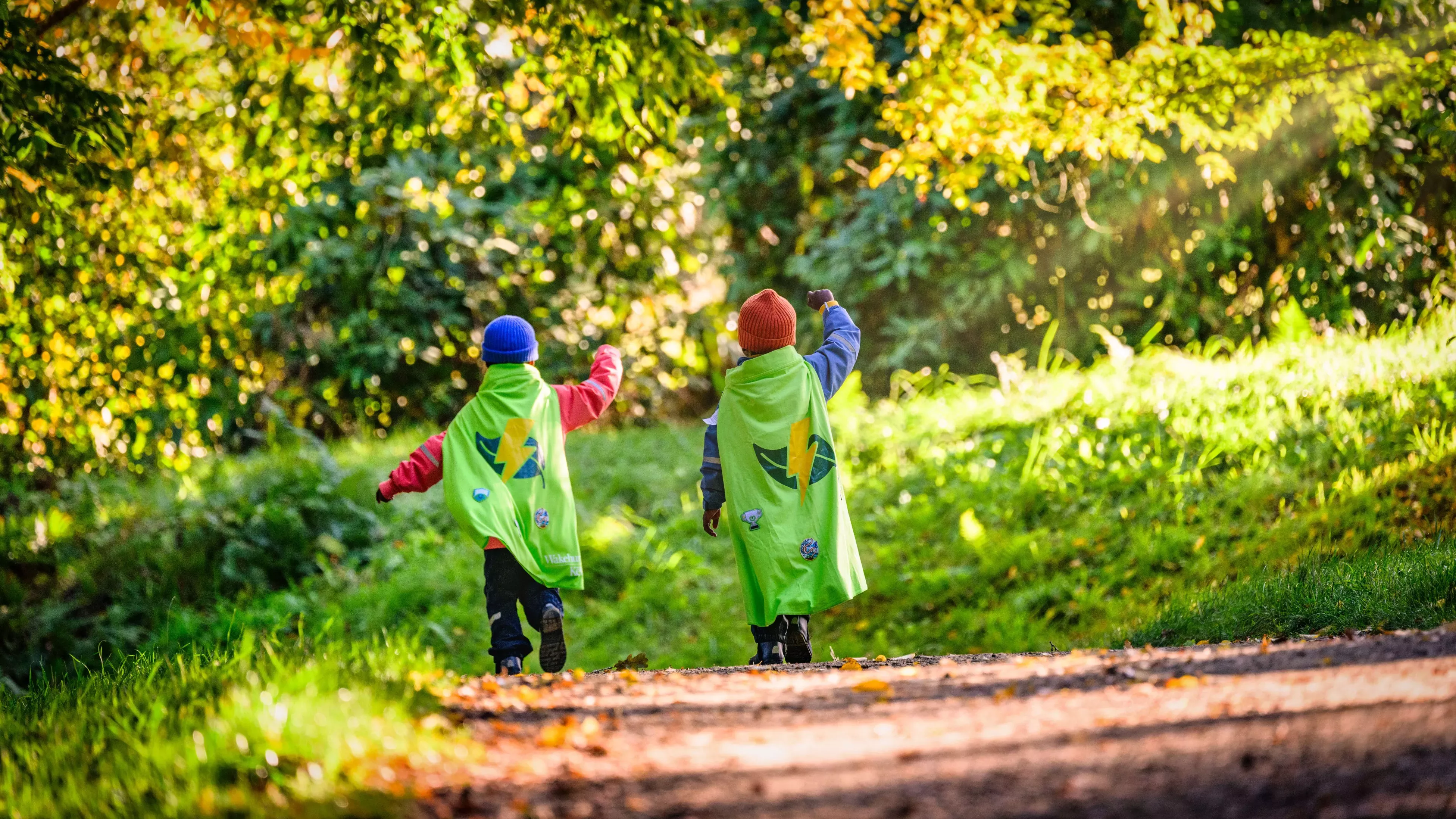 Seen from behind, two children run down a sunny woodland path in autumn, wearing red and blue beanies and green Nature Heroes capes