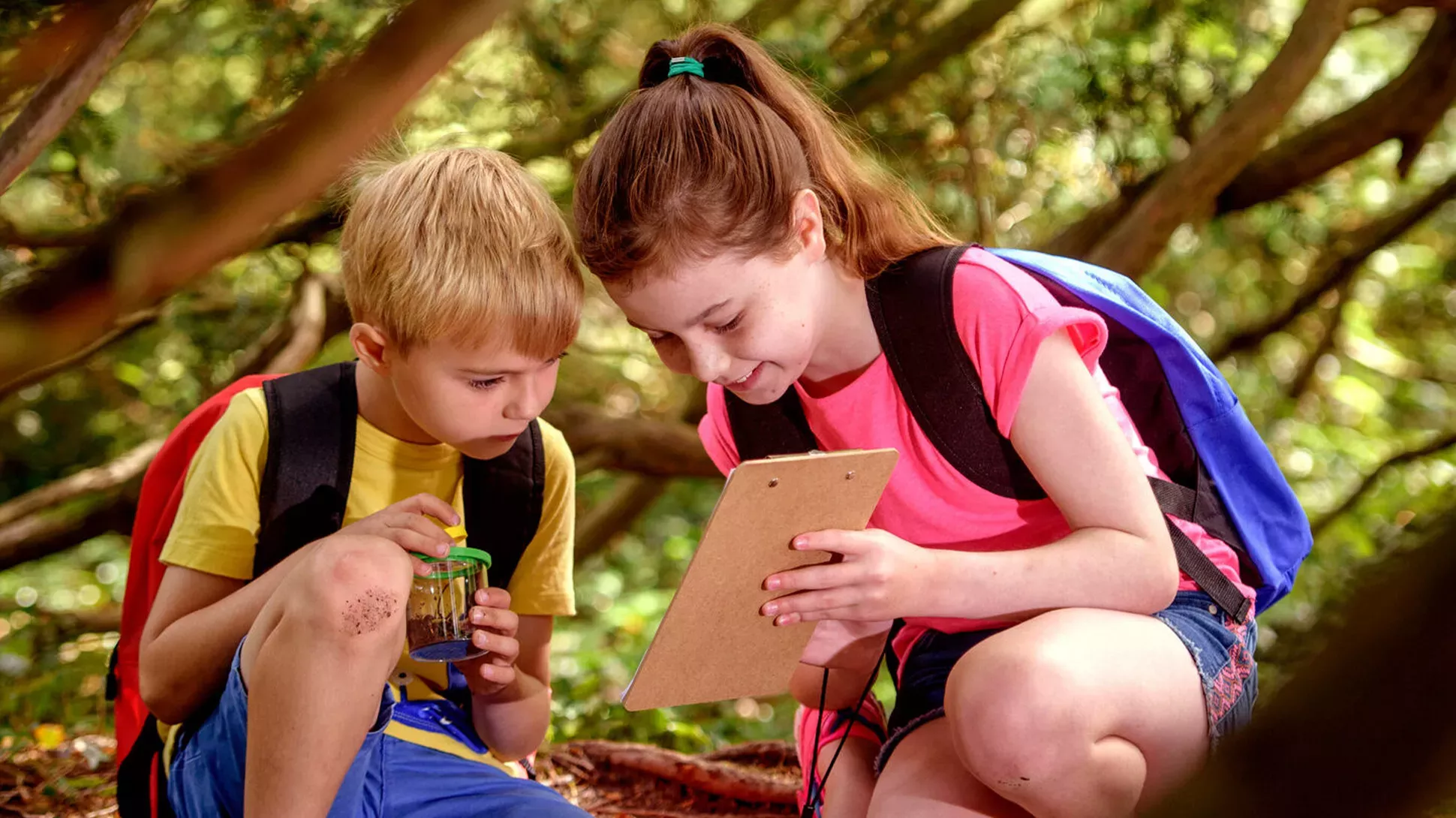 Explorer Rucksacks, Jim Holden © RBG Kew Two children wearing Wakehurst Explorer Rucksacks sit under a tree, looking at a clipboard.