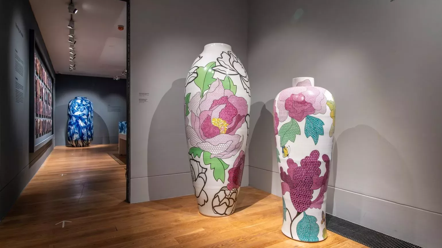 Two large colourful pots in the corner of a gallery room, with a glimpse into the next room as well.